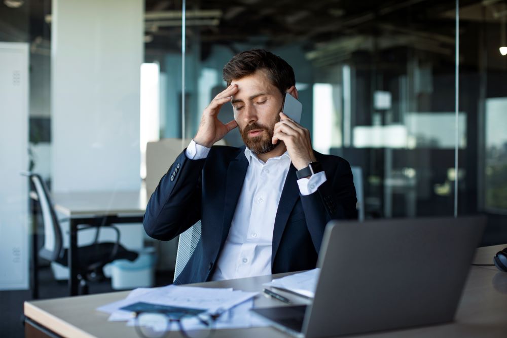 A well-dressed man working at his desk while on a call and looking stressed. Staying grounded: an antidote to our fast digital world.