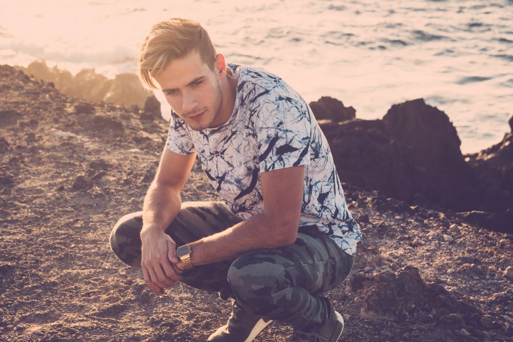 Young attractive man crouched down on a cliff near an Ocean and thinking deeply. Staying grounded: an antidote to our fast digital world.