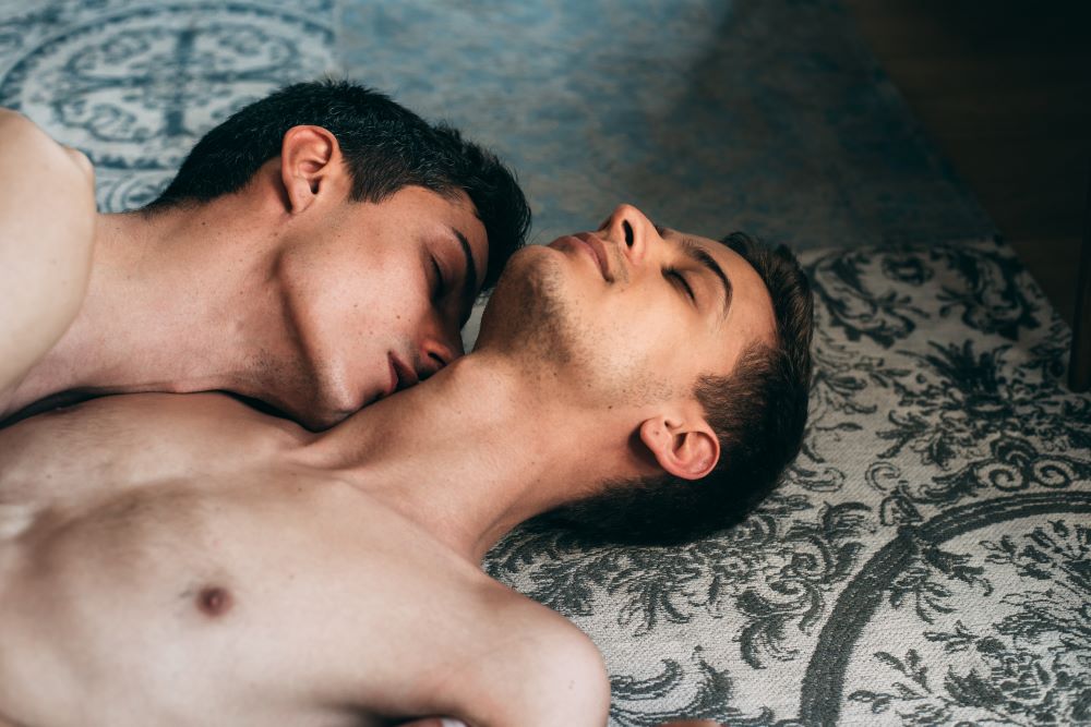 Two men laying intimately together and exploring neck kissing as one of the 5 erogenous zones to make him melt.