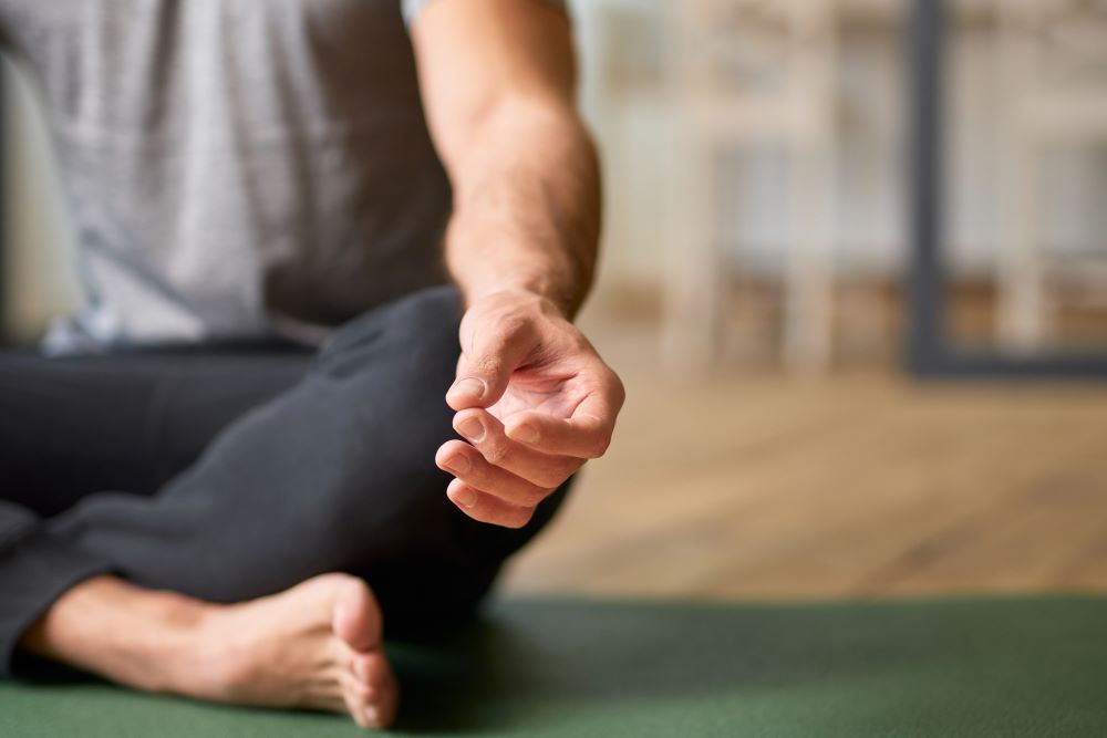 Close-up shot of the torso, forearm, and hand of a young man sitting on the floor with his legs crossed in meditation. Staying grounded: an antidote to our fast digital world.