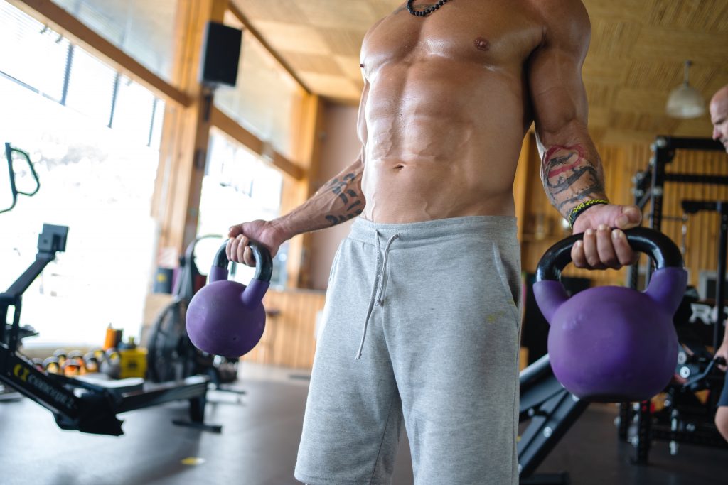 Cropped photo of a fit shirtless man lifting two kettle balls in a gym setting. Pump Up Your Male Sexual Energy With Weight Training