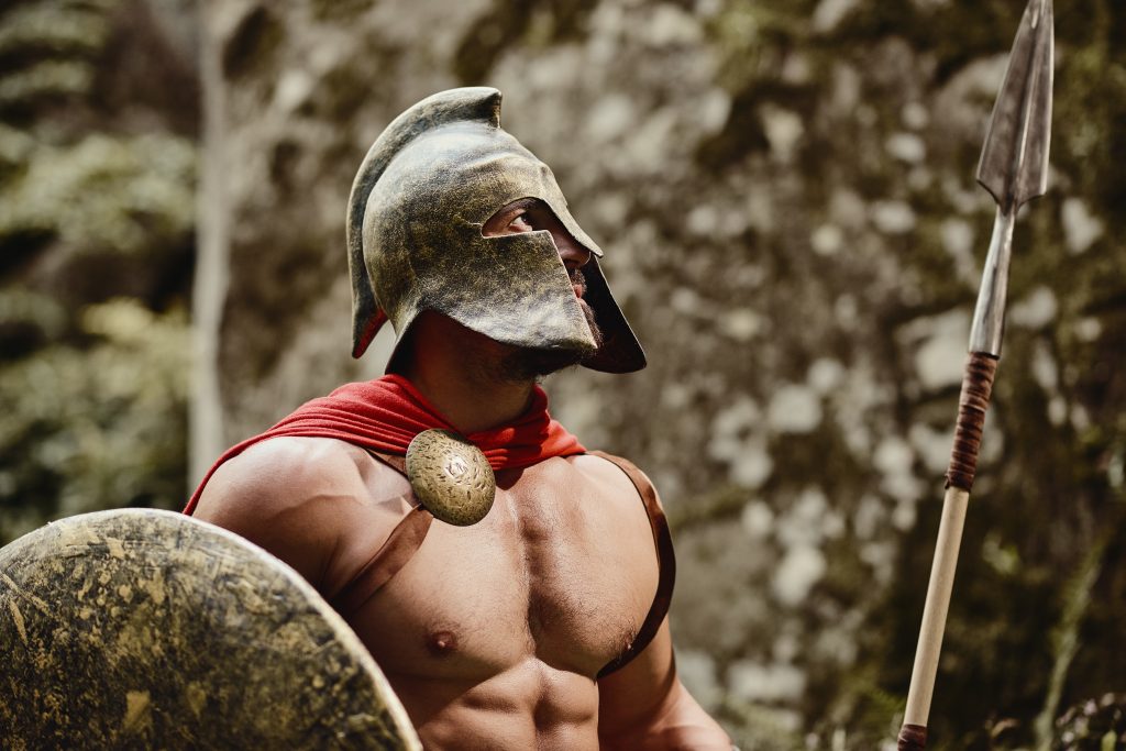 The Not-So Taboo Culture of Gay Relationships and Male Erotic Massage in Ancient Rome. Portrait of anonymous muscular man wearing outfit of ancient gladiator posing in helmet with shield and spear in hands looking away. Spartan.