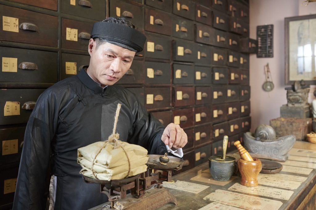 Serious traditional Asian apothecary worker putting plummet on scales to weight package with treatment. Deep Healing from the Transformative Power of Nude Male Erotic Massage.
